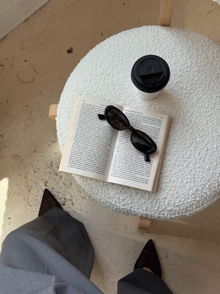 Óculos de sol preto sobre um livro aberto, ao ar livre, com um copo de café